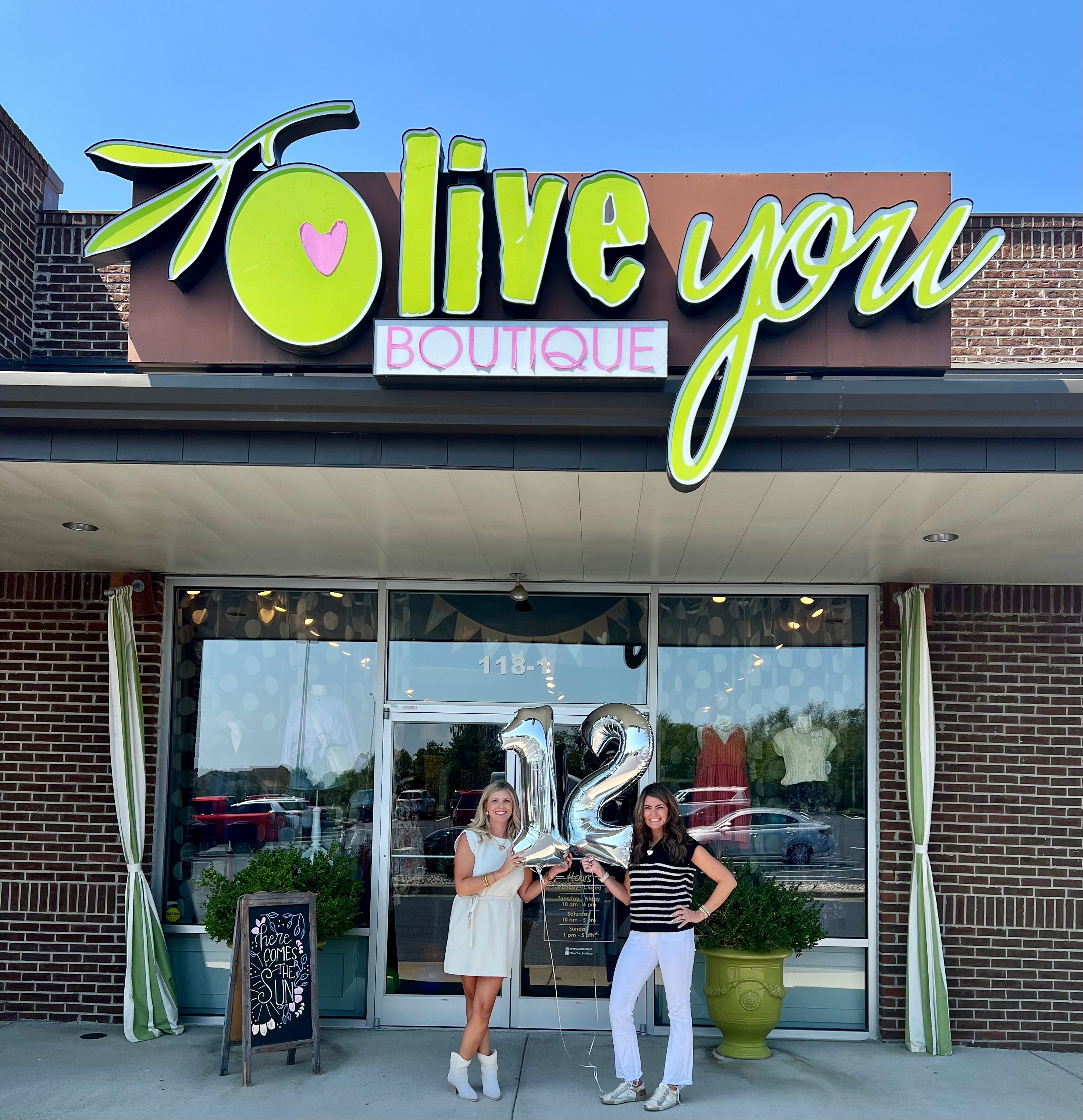 Two women holding a '12' sign in front of 'Olive You Boutique' on a clear day.